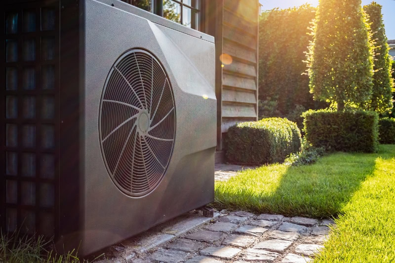 Close-up of black full inverter heat pump outside in the garden, near wooden pool house on a sunny day. Lens flare on the image.