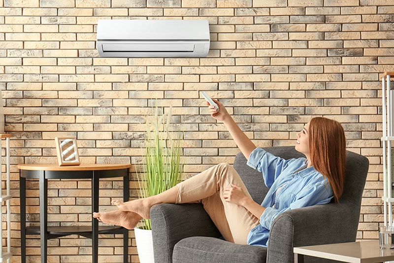 Ductless Mini Splits for Historic Homes. Young woman switching on air conditioner while sitting in armchair at home.